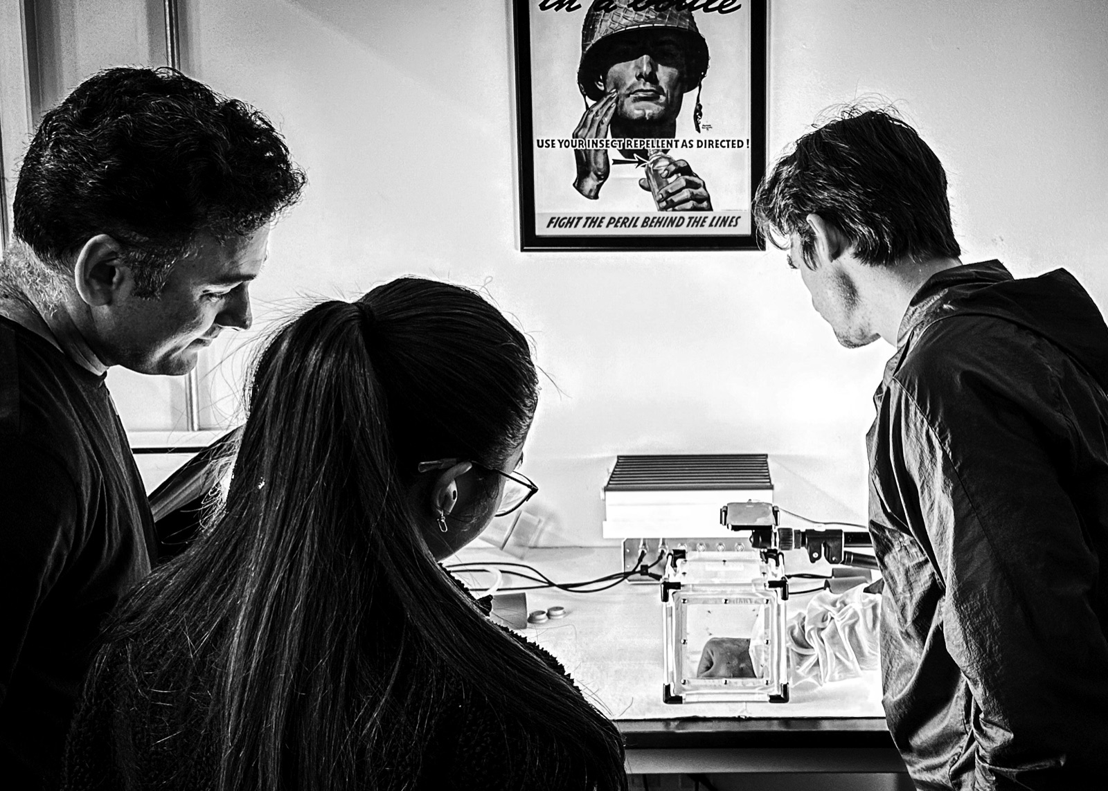 Three researchers at a behavioral assay station, beneath a wartime insect-repellent poster.